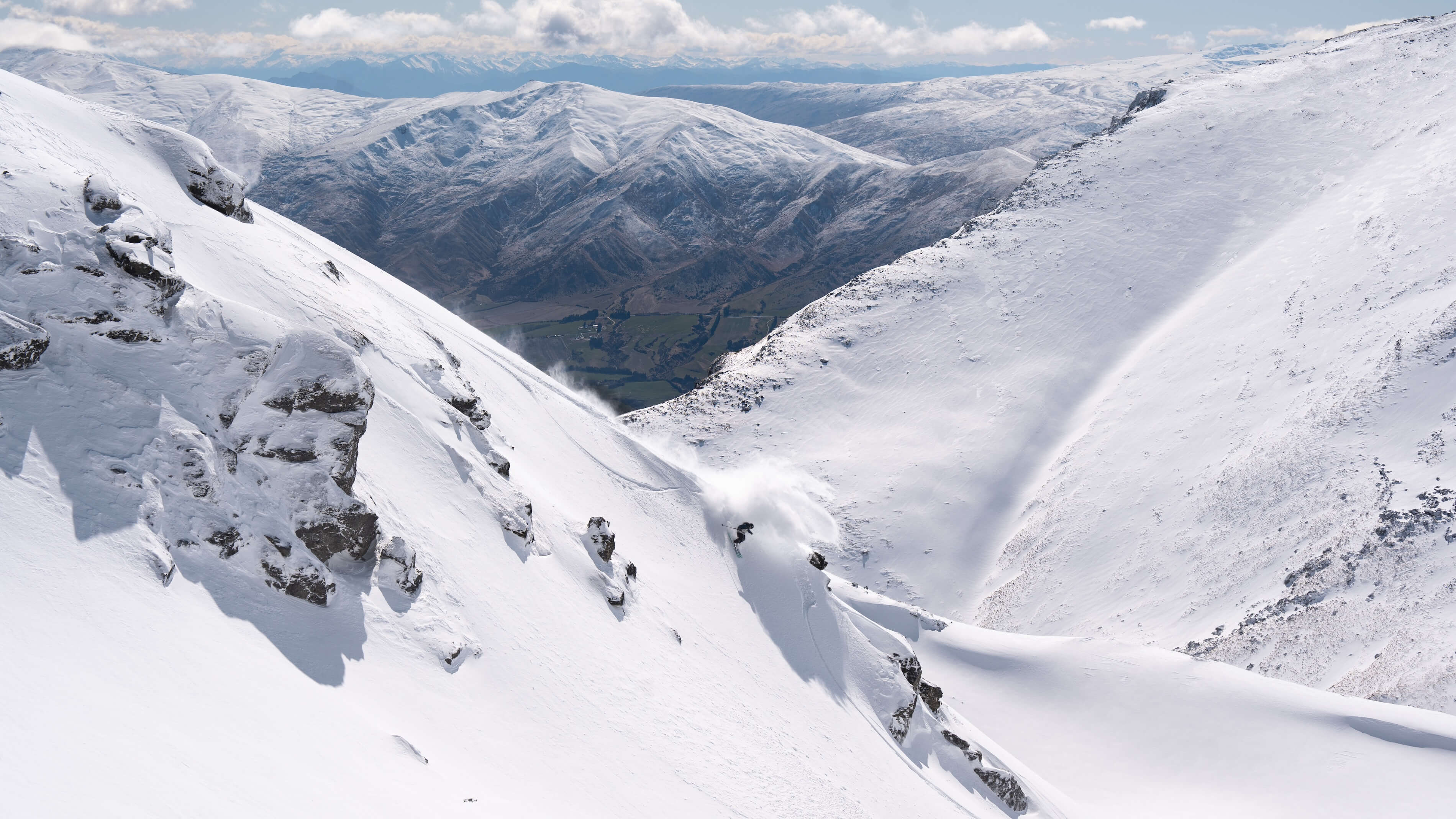 Skier making a sharp turn in deep snow on a snowy mountain slope with rocky outcrops and distant mountains.